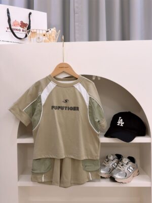 Grey-green T-shirt and shorts on a hanger, with white and green panels and FUFUTIGER lettering on the top and green patch pockets on the shorts; a black cap and trainers sit nearby.