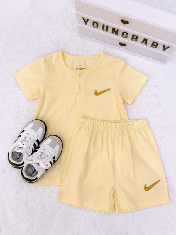 Pale yellow short-sleeved top with two neckline buttons and elastic-waist shorts, featuring golden yellow check-shaped motifs, displayed beside trainers.