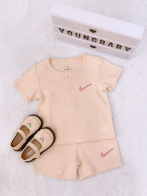 Pale peach round-neck, two-button short-sleeve top and shorts with fine vertical texture and pink tick-shaped motifs, beside beige floral shoes.