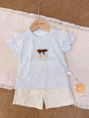 Light heather-grey short-sleeved top with embroidered shoulder ruffles, a brown bow and two round decorations, paired with cream shorts.