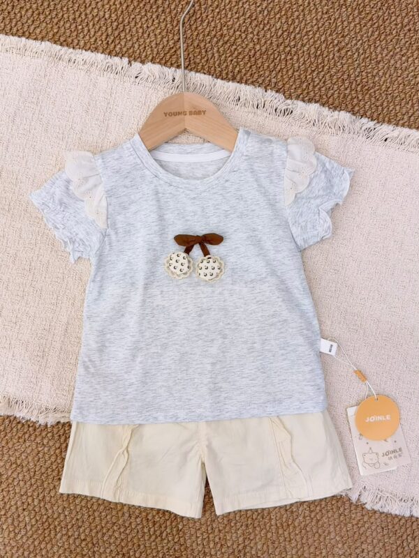 Light heather-grey short-sleeved top with embroidered shoulder ruffles, a brown bow and two round decorations, paired with cream shorts.