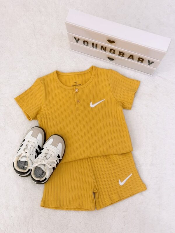 Mustard yellow round-neck, two-button short-sleeve top and shorts with vertical striped texture and white tick-shaped motifs, beside lace-up trainers.