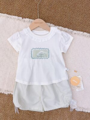 White short-sleeved top with a ruffled round neckline, green rectangular motif and white lace bow, paired with pale grey-and-white gingham shorts with hanging ties at the leg sides.