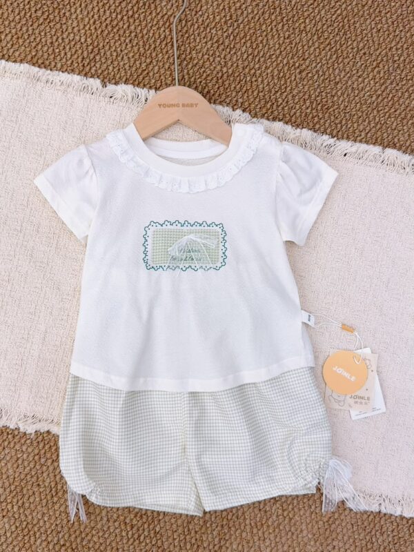 White short-sleeved top with a ruffled round neckline, green rectangular motif and white lace bow, paired with pale grey-and-white gingham shorts with hanging ties at the leg sides.