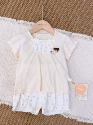 Pale cream short-sleeved top with a gathered white floral neckline panel and small bows, paired with white gathered-hem shorts printed with tiny flowers and cartoon motifs.