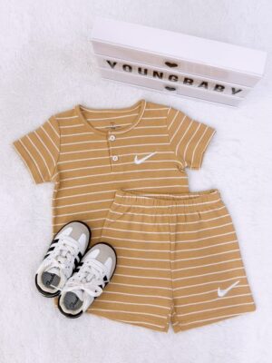 Mustard yellow top and elastic-waist shorts with thin white stripes and white tick-shaped motifs, laid flat beside trainers and a YOUNGBABY lightbox. The short-sleeve top has a round neck and button placket.