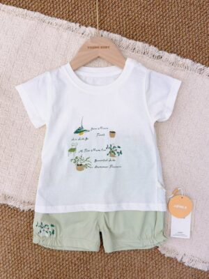 White crew-neck short-sleeved top with green plant, pendant lamp and chair graphics, paired with pale green shorts with a plant motif near the hem.