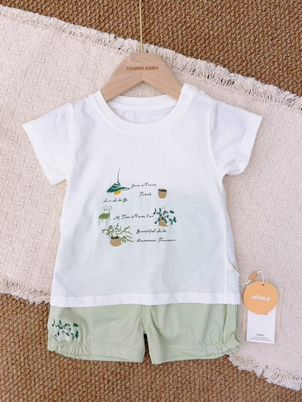White crew-neck short-sleeved top with green plant, pendant lamp and chair graphics, paired with pale green shorts with a plant motif near the hem.